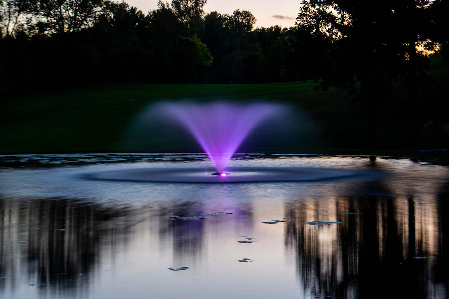 Kasco 5 HP display fountain pond aerator 240V spraying purple-lit fan pattern on large pond at dusk